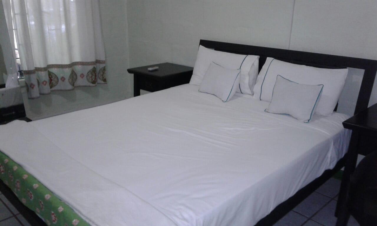 A neatly made bed with white linens and pillows, a small bedside table, and a curtain with a decorative border.