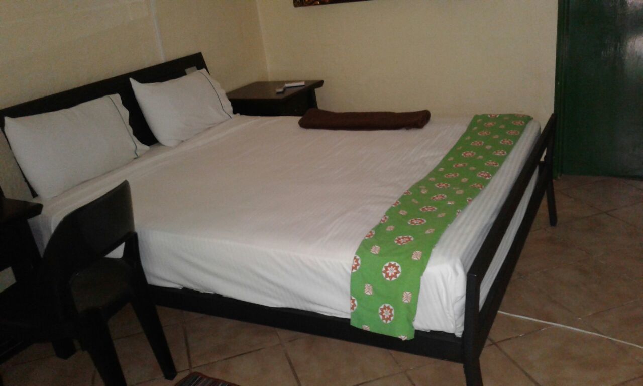 A single bed with a white mattress and a green patterned bedspread. There is a brown pillow and a remote control on a small bedside table. The room has a tiled floor and a green door.