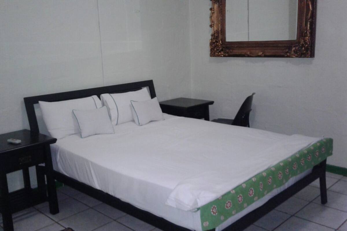 A neatly made bed with white linens, a green patterned bedspread, and a wooden headboard. There are two bedside tables with black frames, one of which has a remote control on it. A mirror with an ornate wooden frame hangs on the wall above the bed.