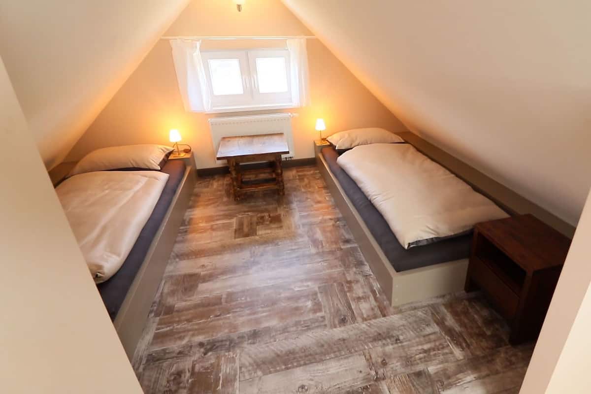 A small, triangular attic bedroom with two single beds, a wooden floor, a window with curtains, and a nightstand with a lamp.