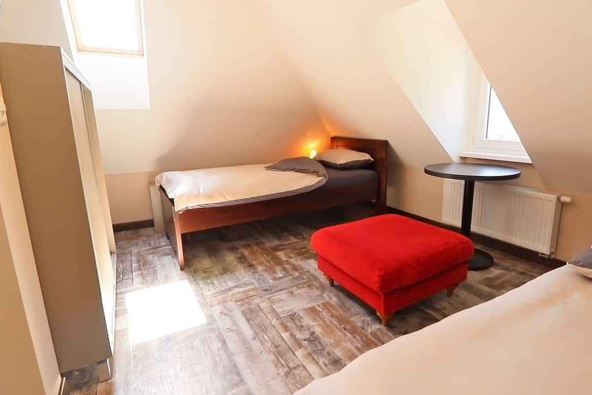 A compact room with a bed, a red ottoman, a small table, and a window.