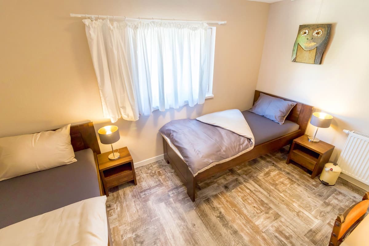 A bedroom with two single beds, a wooden floor, a window with white curtains, and a wall-mounted artwork.