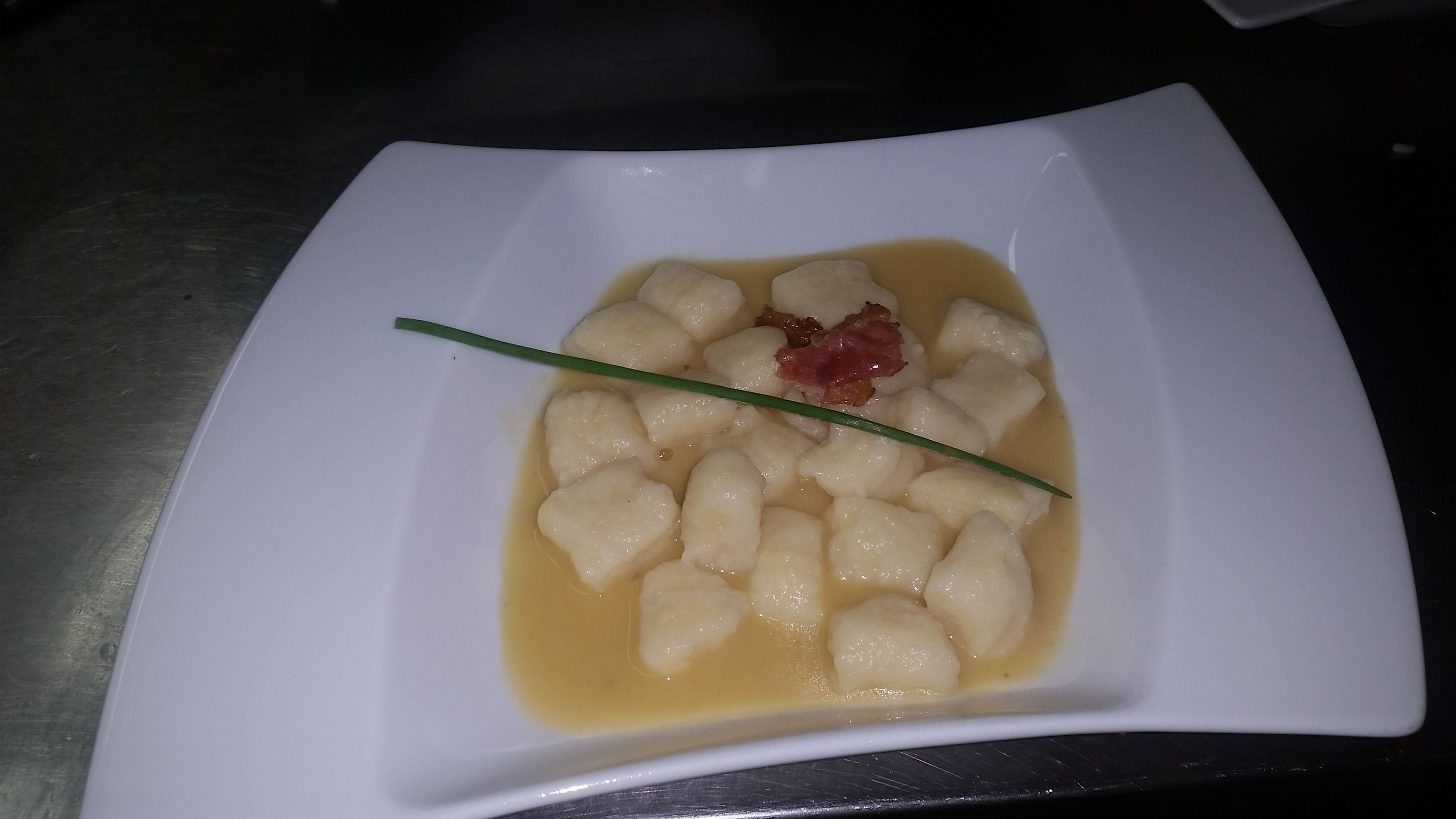 A square white plate with a dish of gnocchi in a light sauce, garnished with a chive and a piece of bacon.