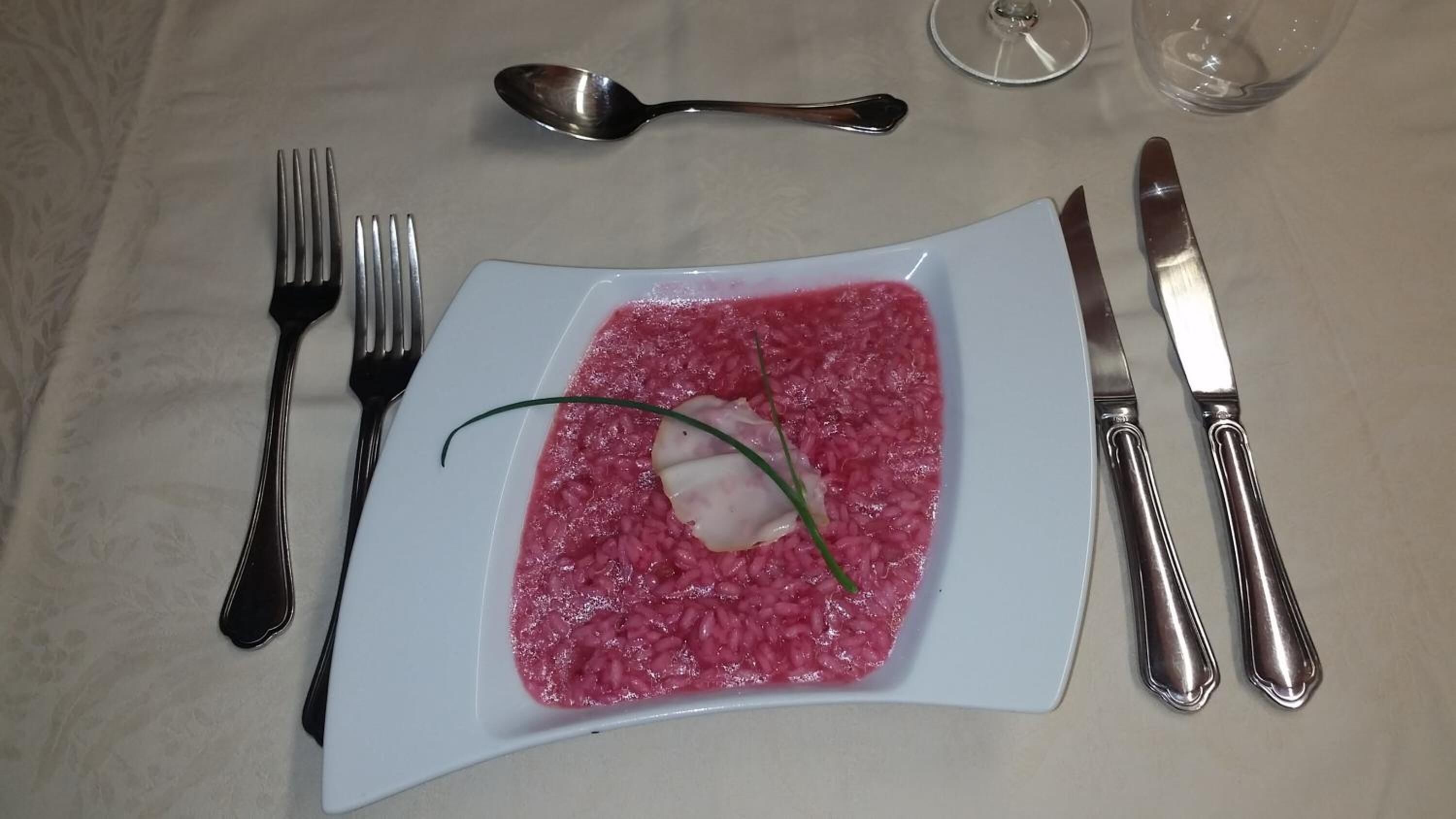 En tallerken rød risotto med pynt, bestik og et glas vand på et bord.