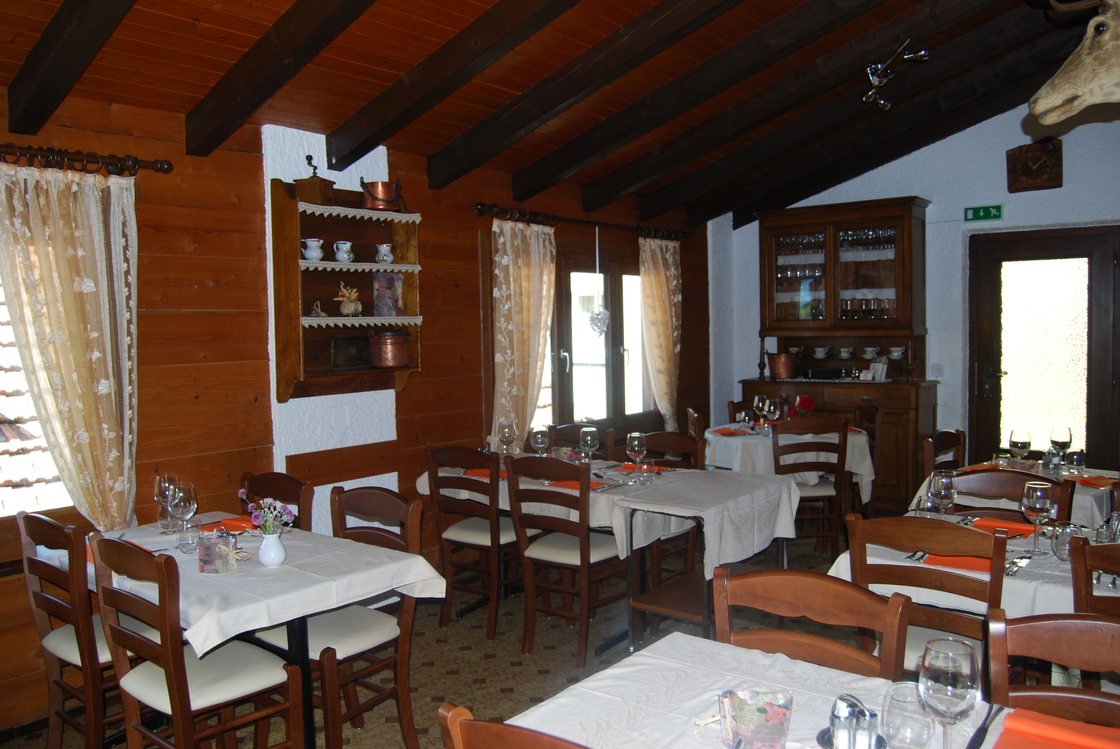 A dining area with wooden tables and chairs, white tablecloths, and a wooden cabinet with glass doors.
