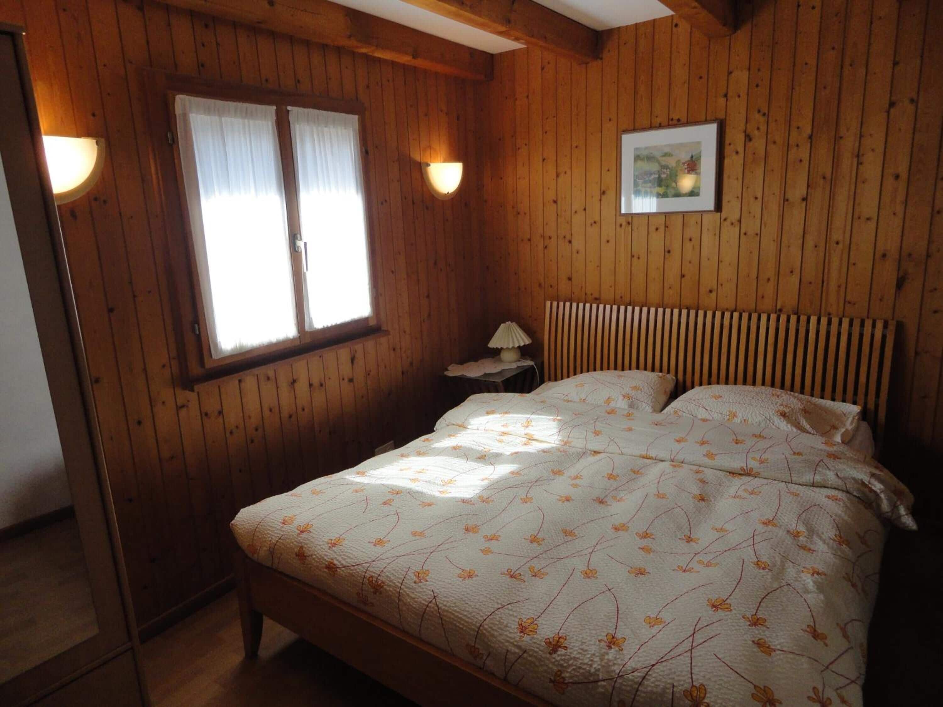Une pièce aux murs lambrissés, comprenant un lit, deux lampes fixées au mur, une fenêtre avec des rideaux et un tableau encadré accroché au mur.