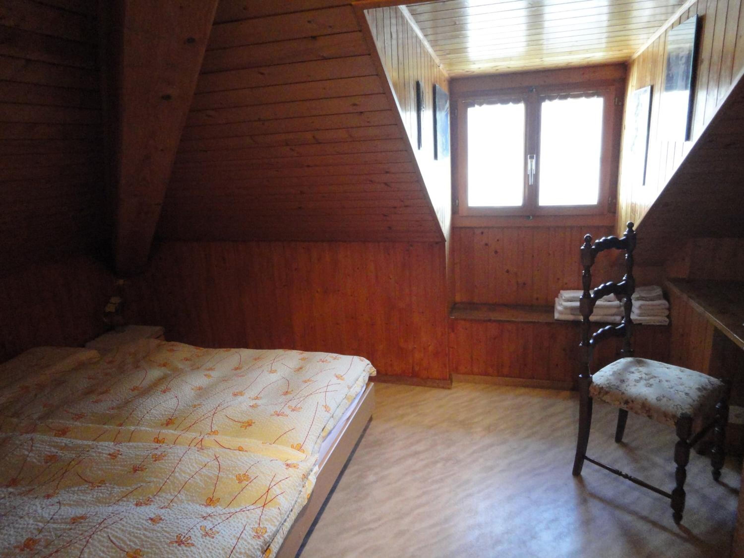 Un dormitorio con techo inclinado de madera, una cama con colcha floral, una silla con cojín floral y una ventana con vista al exterior.