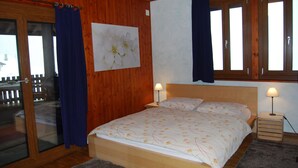 A bedroom with a wooden floor, a bed with floral bedding, two bedside lamps, and a window with curtains. - Osteria Anzonico (Faido)