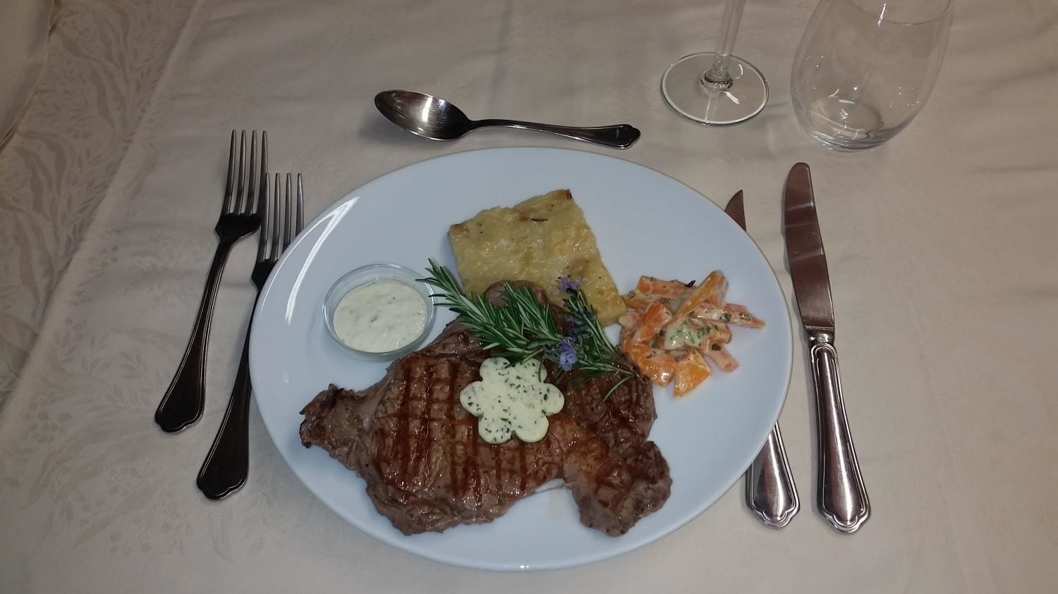 Ein Teller mit einem Steak, einem Stück Brot und einem Beilagensalat, dazu Gabel, Messer und Löffel auf einer Tischdecke.