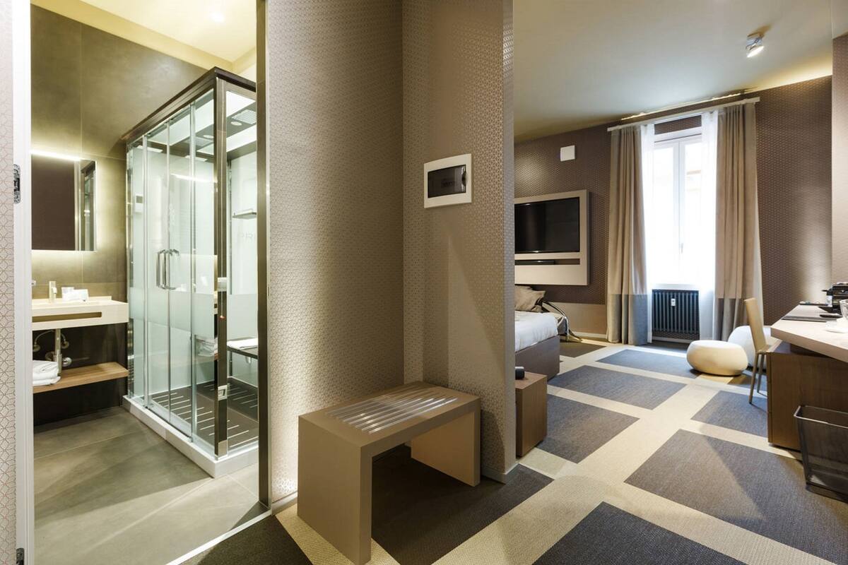 A modern hotel room with a glass shower enclosure, a bed with a TV, a small seating area, and a large window with curtains.