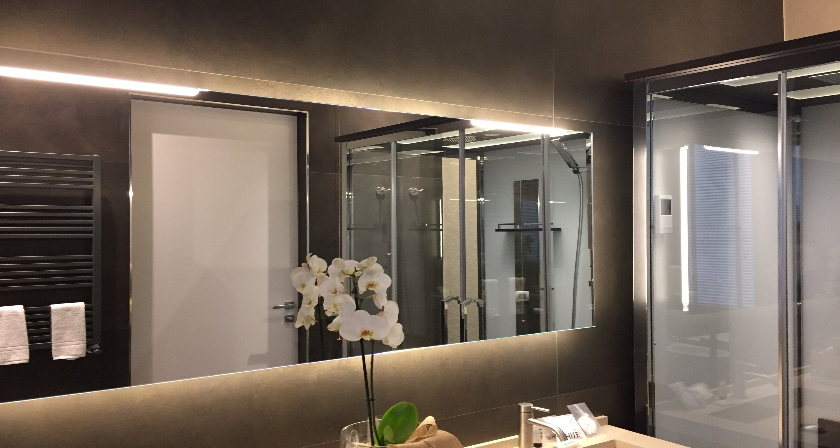 A modern bathroom with a large mirror, a glass shower enclosure, and a vase with white flowers on the sink.
