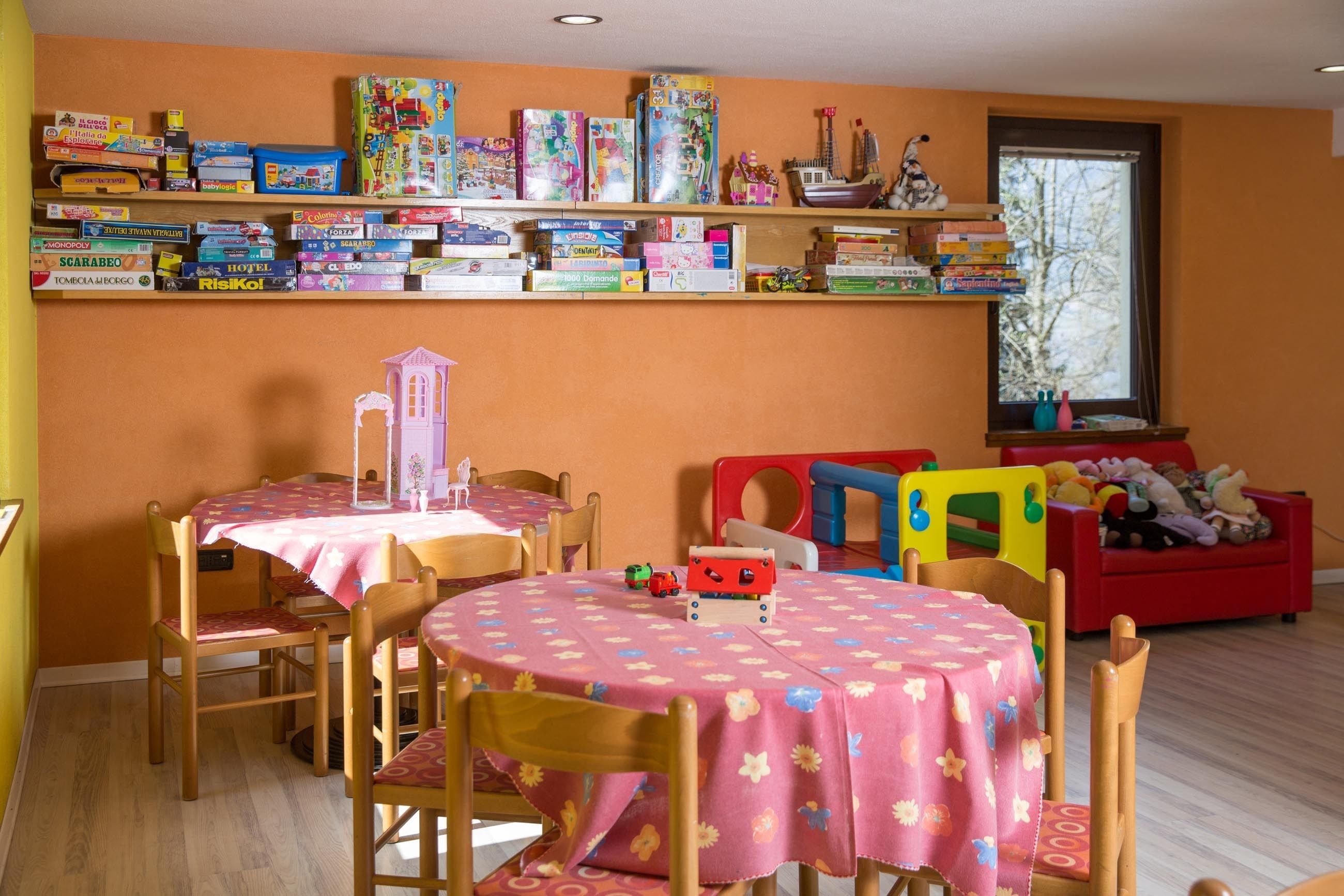 A children's playroom with a round table covered in a pink floral tablecloth, surrounded by wooden chairs. Above the table, there is a shelf with various board games and toys.