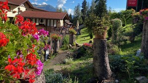 Un hotel con un edificio de estilo chalete tradicional, un jardín con diversas plantas y flores, y un paisaje montañoso de fondo.