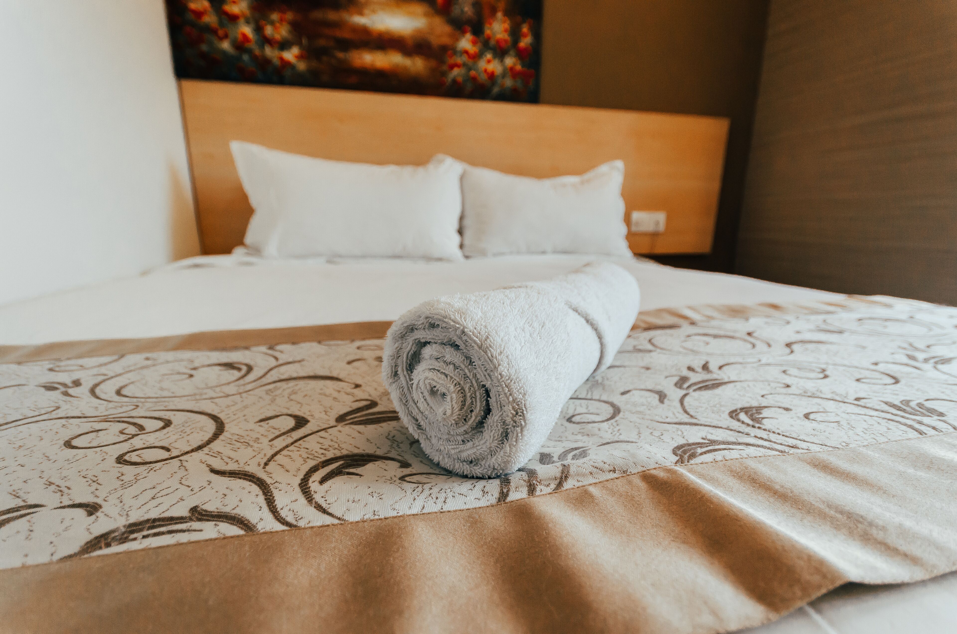 A neatly made bed with a rolled towel, a patterned bedspread, and a painting on the wall.