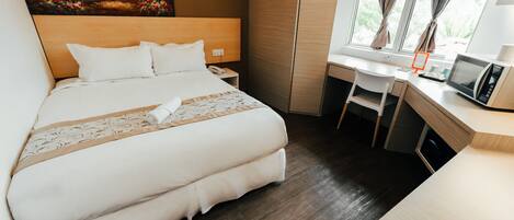 Una cama bien tendida con sábanas blancas y una almohada decorativa, un escritorio de madera con una computadora y un teléfono, y un cuadro en la pared.