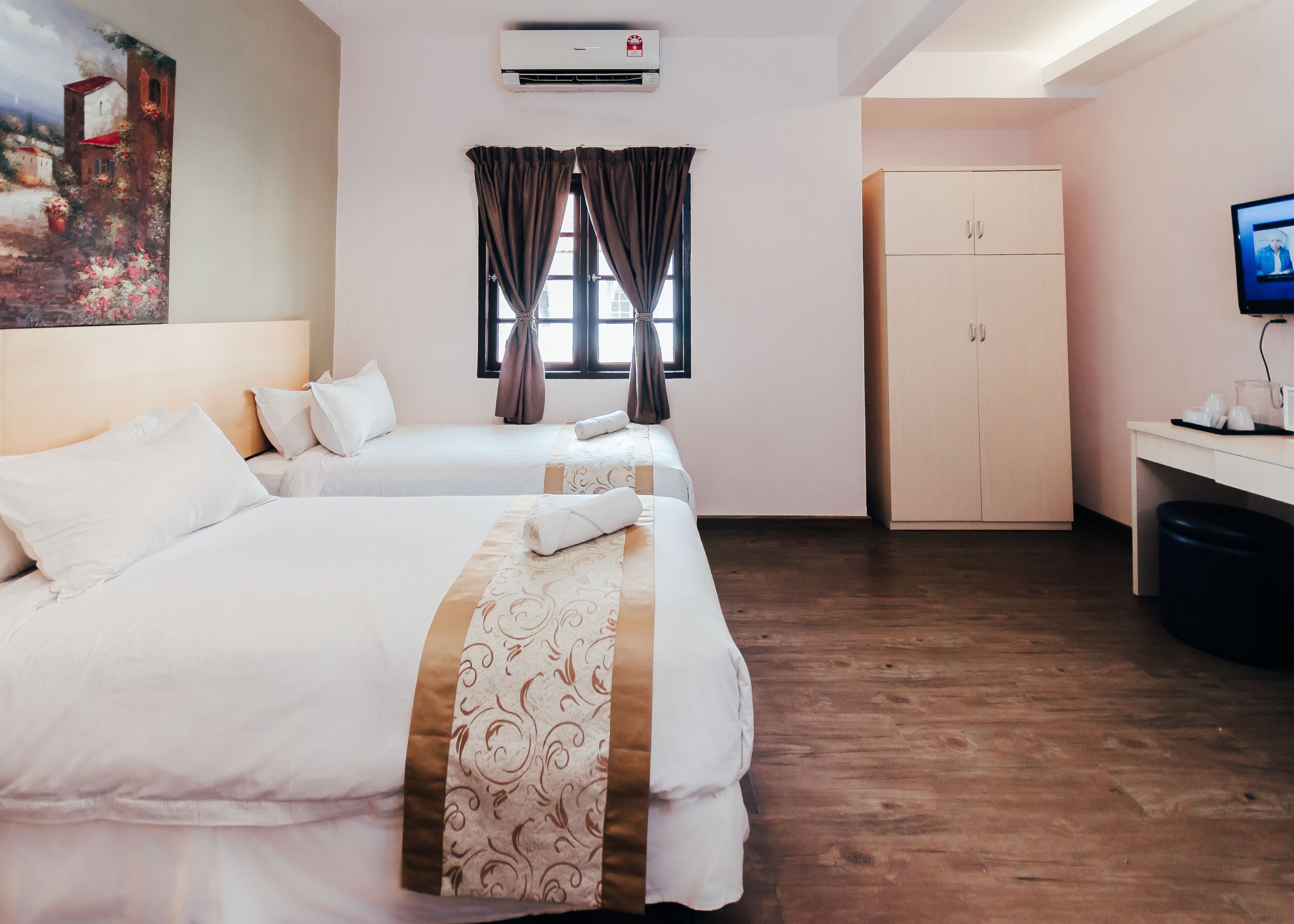 A hotel room with two beds, a wardrobe, a desk with a computer, and a painting on the wall.
