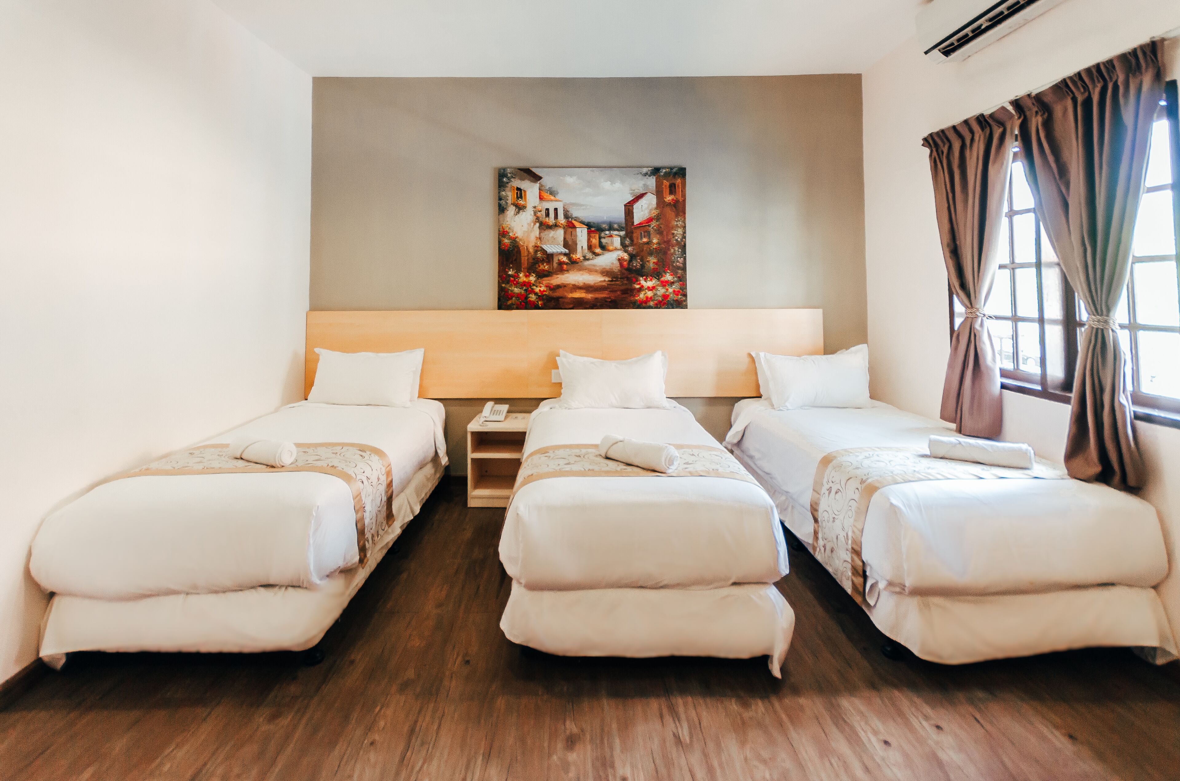 A hotel room with two single beds, a wooden nightstand, a painting on the wall, and a window with curtains.