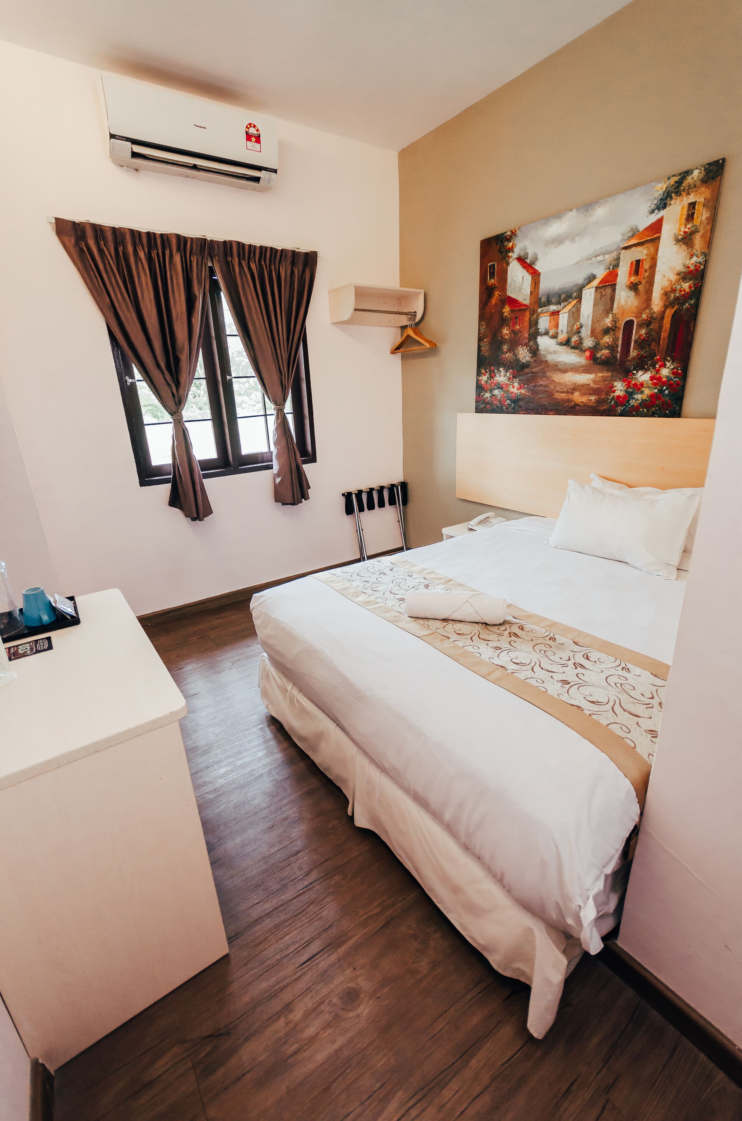 Quarto de hotel com cama, escrivaninha, ar condicionado, janela com cortinas e um quadro na parede.