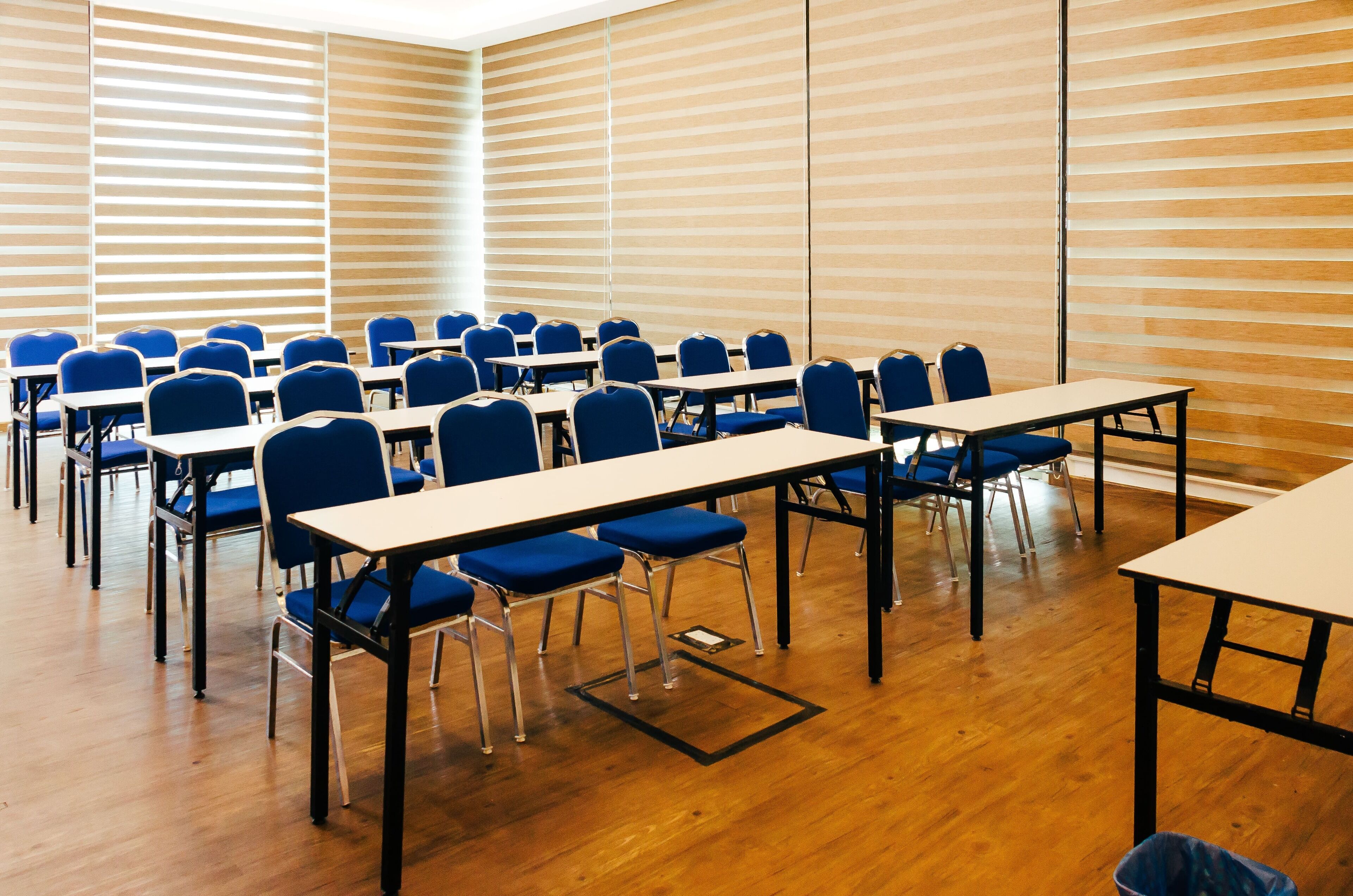 Une salle de conférence avec des rangées de chaises et de tables bleues, un plancher en bois et de grandes fenêtres équipées de stores.