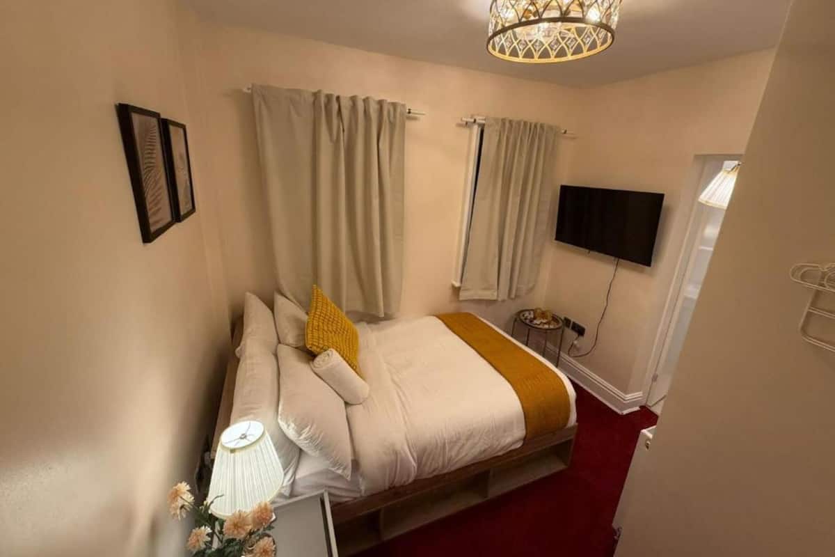 Un dormitorio con cama, cortinas, televisor, lámpara y una pequeña mesa con flores.