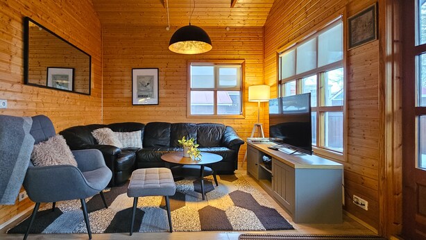Un interior de cabaña acogedor, con paredes de madera, un sofá de cuero, una mesita con una lámpara y un televisor.
