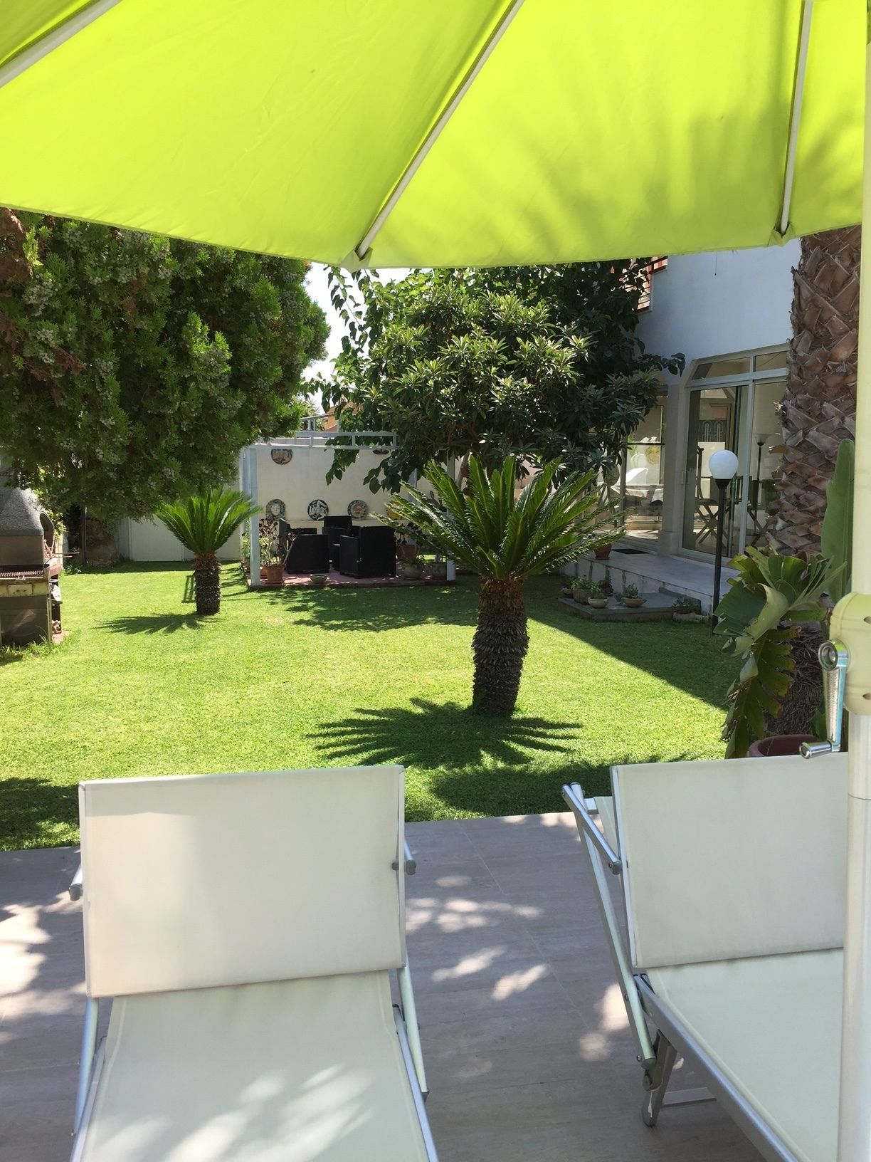 An outdoor patio area with a green umbrella, white lounge chairs, and a small garden with palm trees and a white RV in the background.