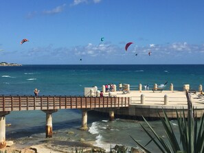 A pier with a wooden walkway over water, people sitting and standing, and kites flying in the background.