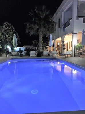 A swimming pool with a blue tint, surrounded by lounge chairs and umbrellas, with a building in the background.