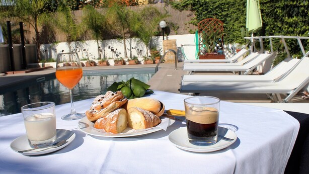 A table with a plate of pastries, a glass of milk, and a cup of coffee, overlooking a pool area with lounge chairs and a view of buildings and greenery. - B&B Marricriu (Avola)
