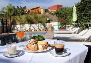Una mesa con un plato de pasteles, un vaso de leche y una taza de café, con vistas a una zona de piscina con sillas de descanso y vistas a edificios y vegetación.