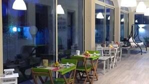 A dining area with green chairs and white tables, large windows, and hanging pendant lights. - B&B Marricriu (Avola)
