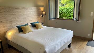 A bedroom with a large bed, a wooden headboard, a window with a view of greenery, and a wall-mounted light fixture.