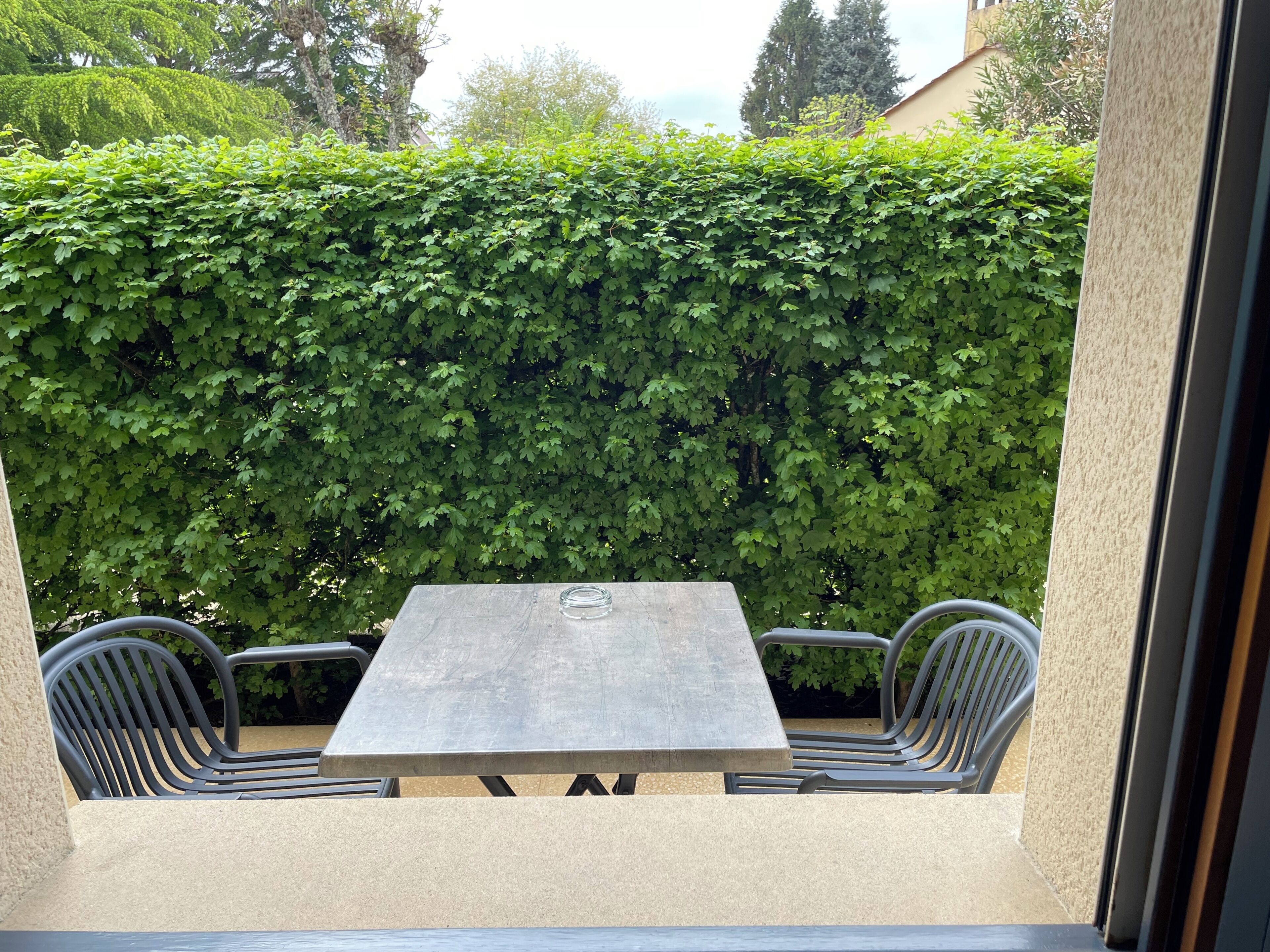 En uteplats med ett bord och två stolar, en häck i bakgrunden och en byggnadsfasad.