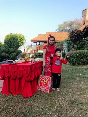 Una persona de pie junto a una mesa con mantel rojo, una caja decorativa y un edificio con tejado de tejas al fondo.