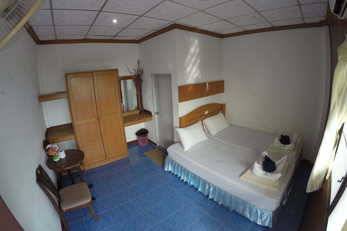 Ein Hotelzimmer mit einem Bett, einem Holzschrank, einem Schreibtisch mit Stuhl, einem kleinen Tisch mit Blumenvase und einem Deckenventilator.