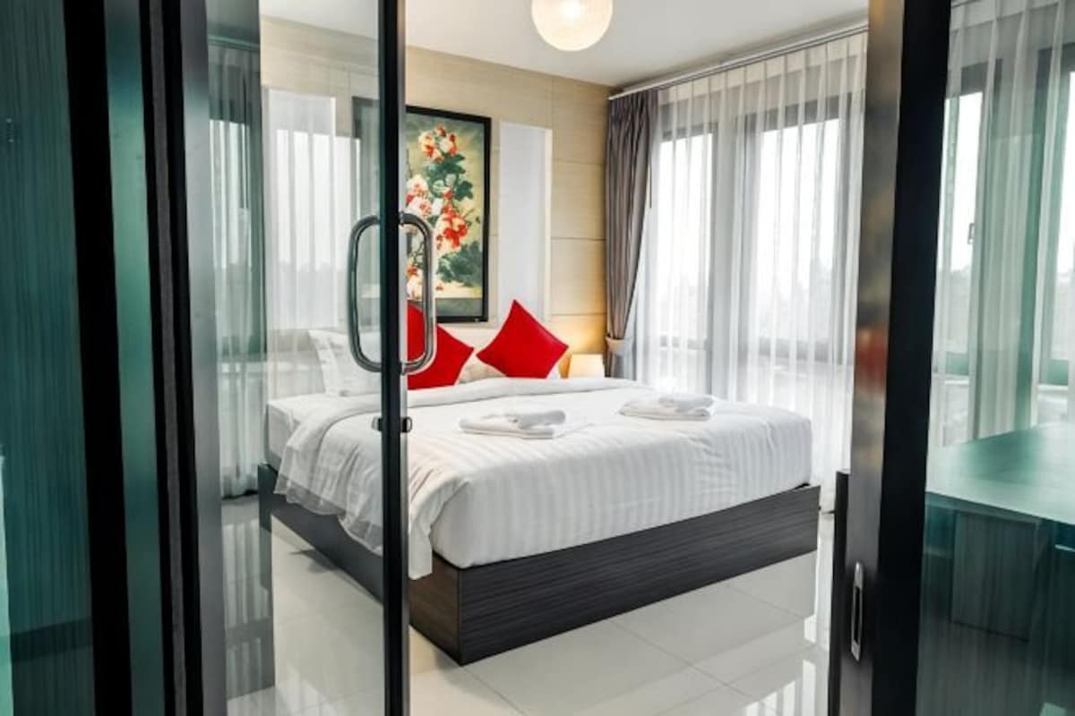 A modern hotel room with a large bed, a glass door, and a view of the city.