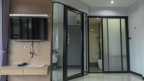 Une chambre d’hôtel moderne dotée d’un téléviseur à écran plat fixé au-dessus d’une console en bois, d’une porte coulissante en verre donnant sur une salle de bain et d’un sol carrelé, propre et bien entretenu.