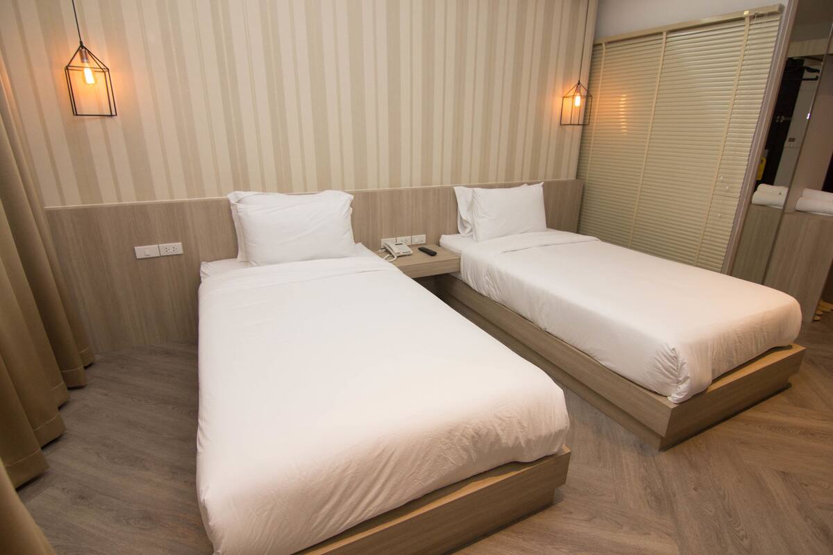 A hotel room with two single beds, a wooden headboard, a nightstand with a phone, and a window with blinds.