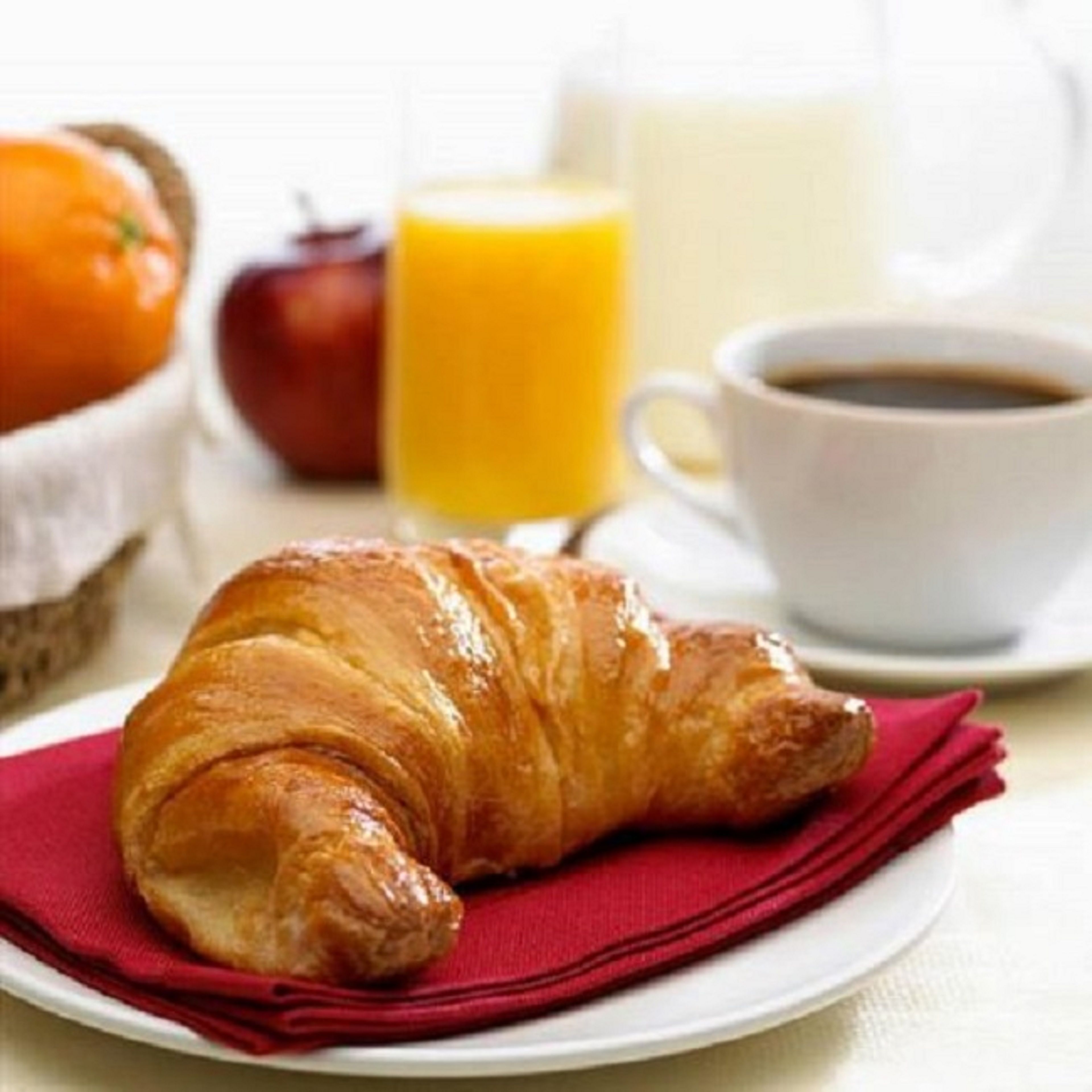 Ein Croissant auf einem Teller mit einer roten Serviette, ein Glas Orangensaft, eine Tasse Kaffee und ein Korb mit einem Apfel und einer Tomate.