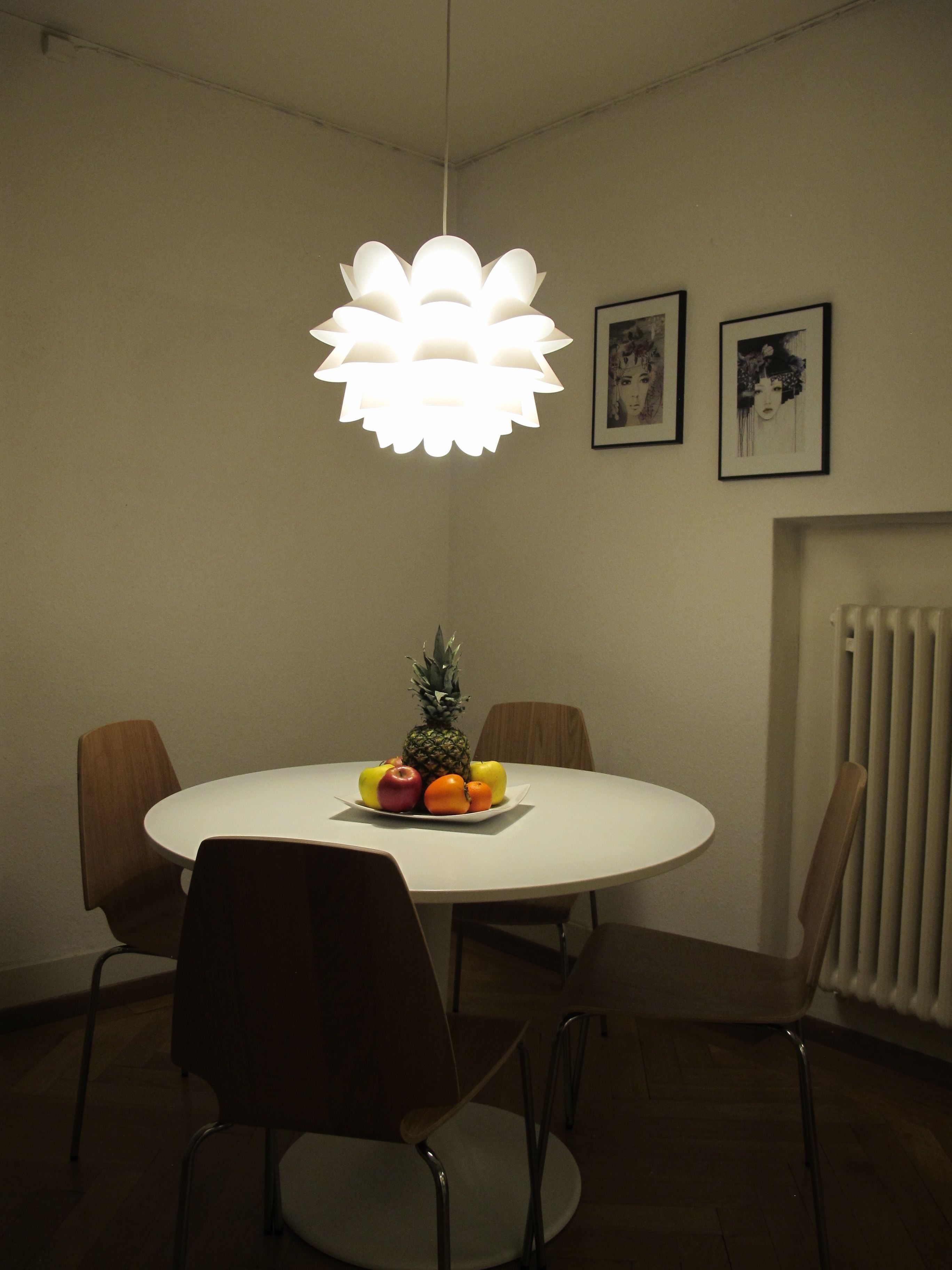 A round dining table with a white surface, surrounded by four chairs. A pineapple and assorted fruits are placed on the table. A unique, flower-shaped pendant light hangs above.