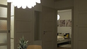 Una habitación con una mesa redonda blanca que presenta una bandeja de frutas con piña, manzanas y una naranja. Sobre la mesa, una lámpara blanca con forma de flor.