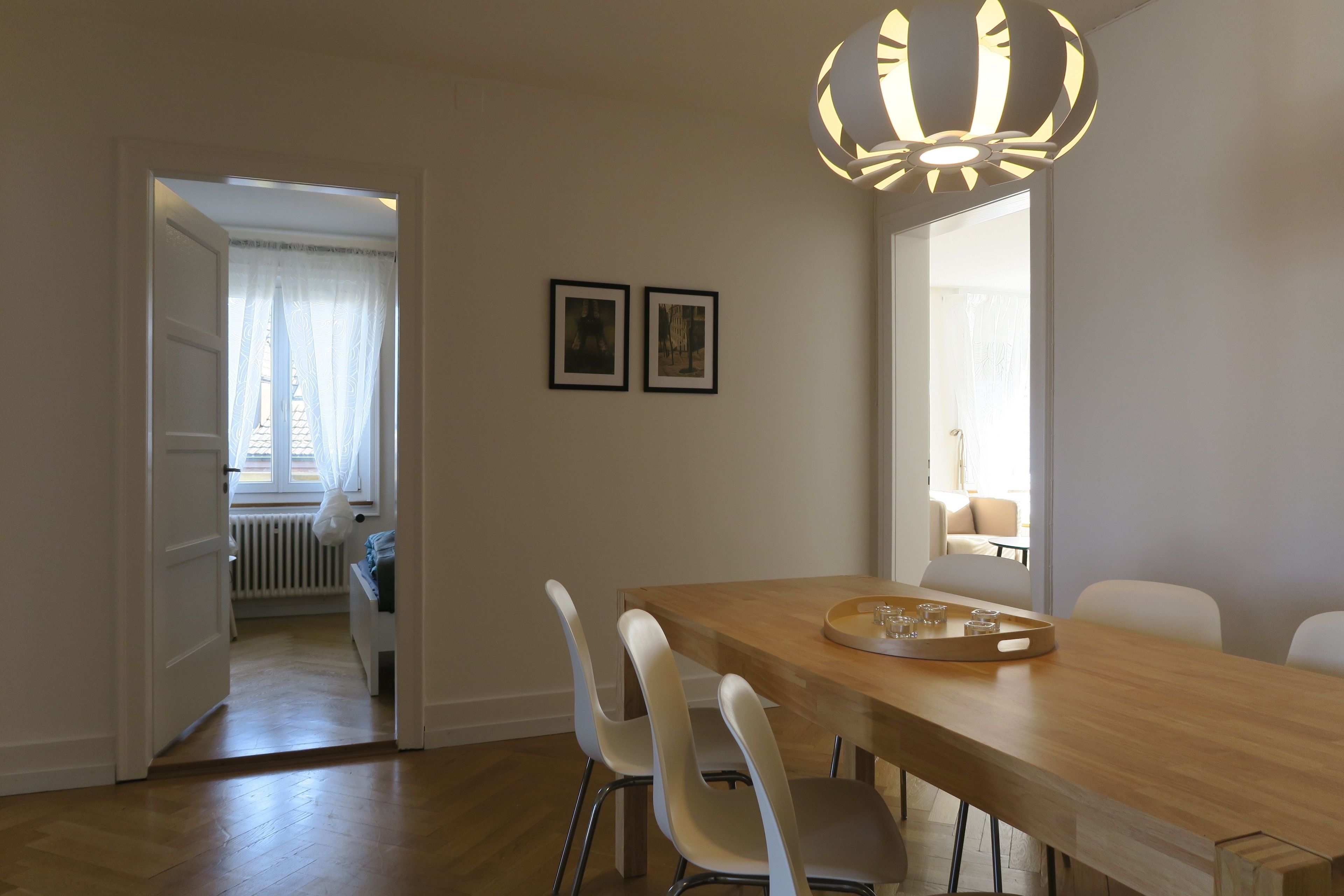 Een eethoek met een houten tafel, witte stoelen en een moderne hanglamp. Er hangen twee afbeeldingen in lijsten aan de muur.