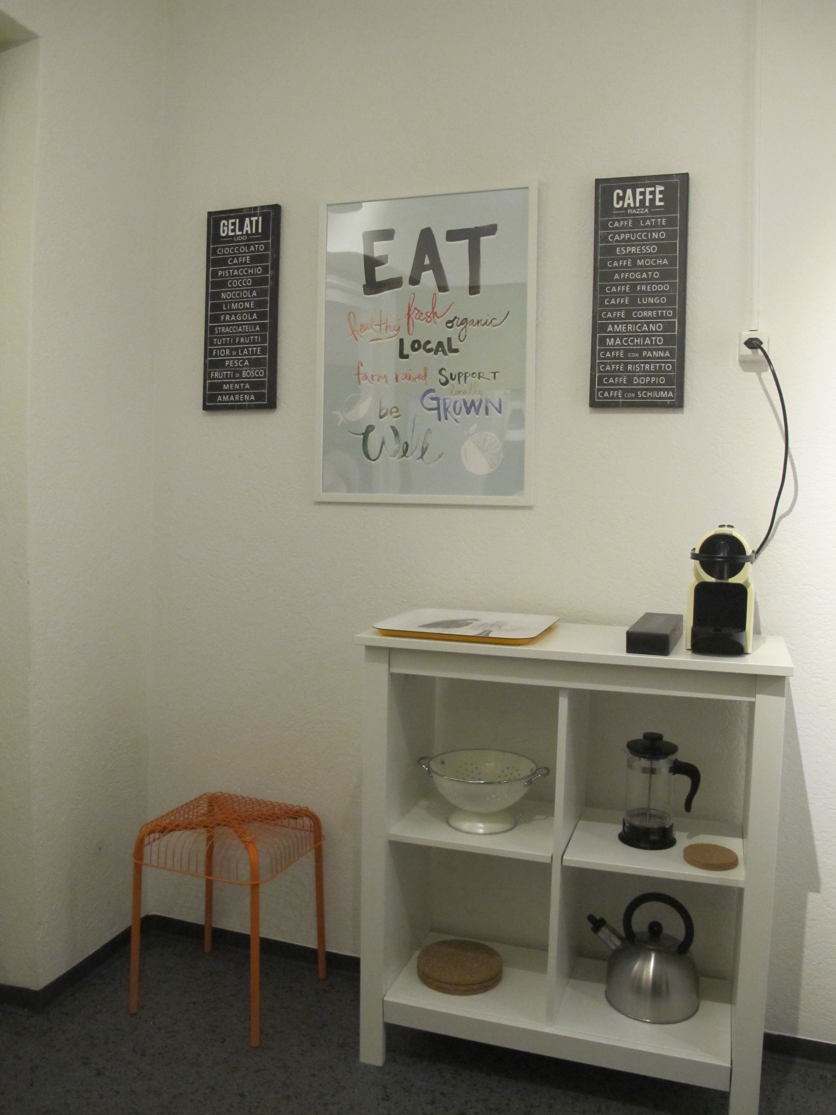 A kitchen area with a coffee machine, a kettle, and a bowl on a shelf. There are two signs on the wall listing food and drink items.