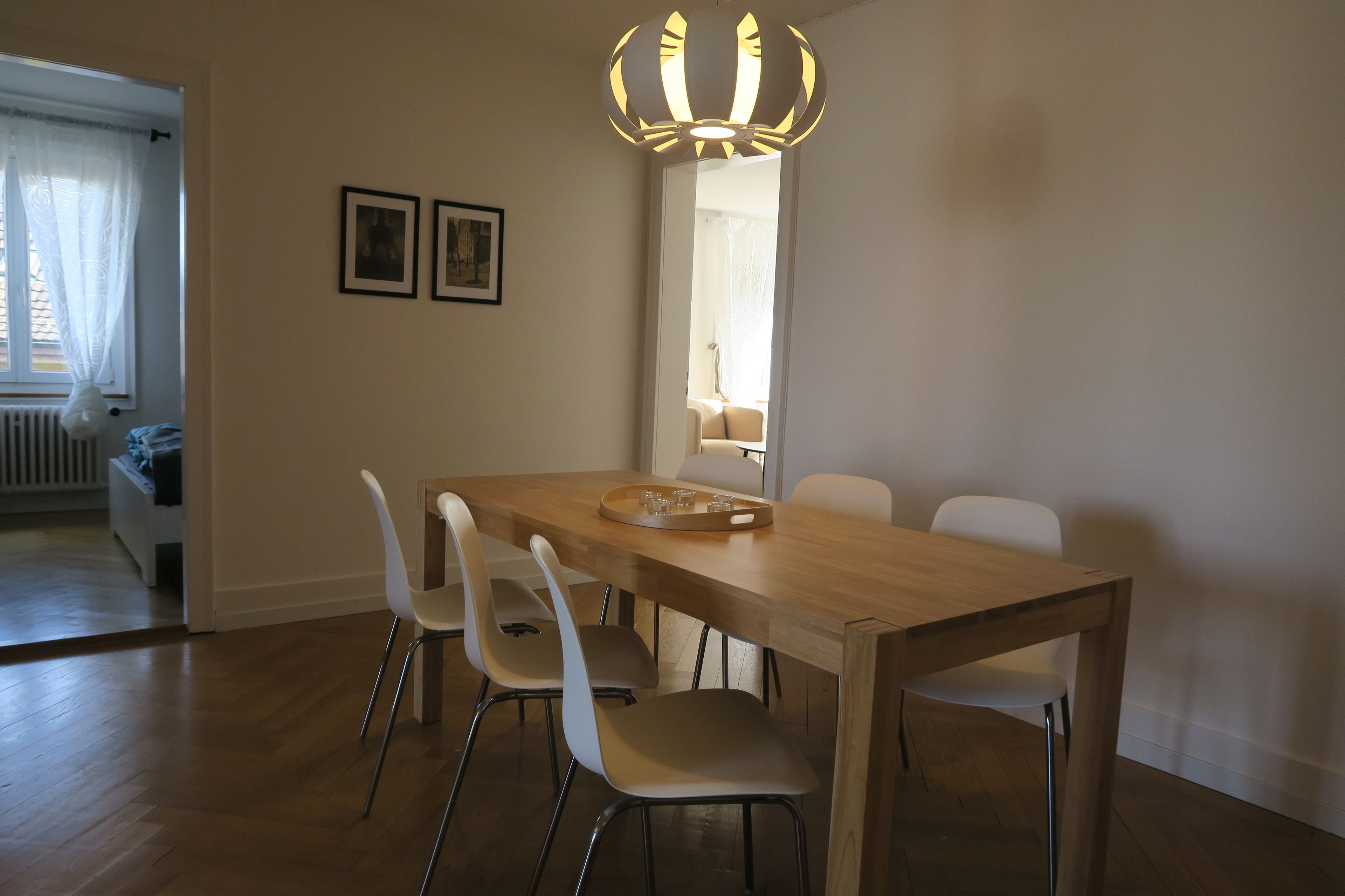 Un comedor con una mesa de madera, cuatro sillas blancas, una araña y dos cuadros en la pared.