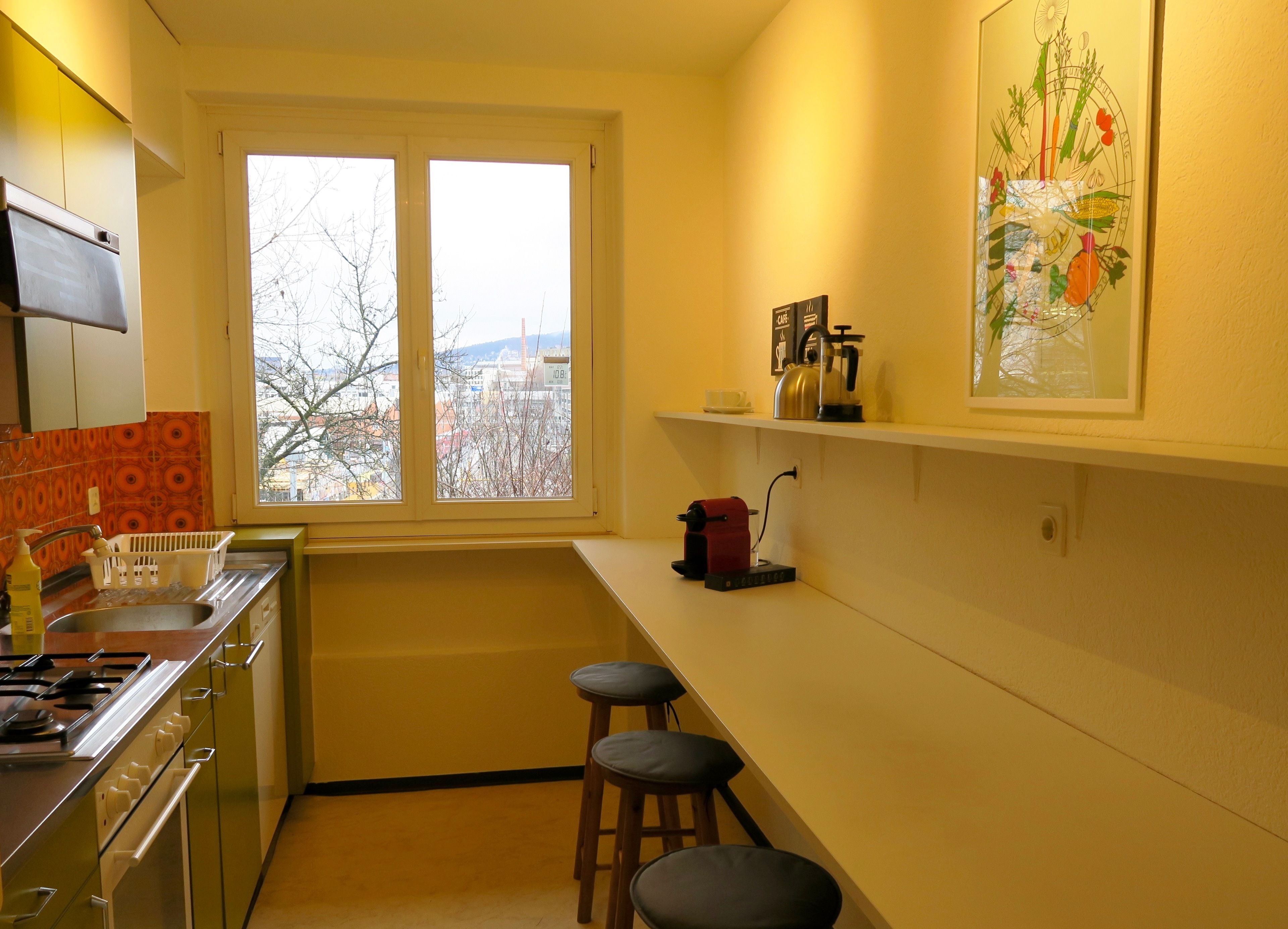 A compact kitchen with a window, a stove, a sink, and two stools.