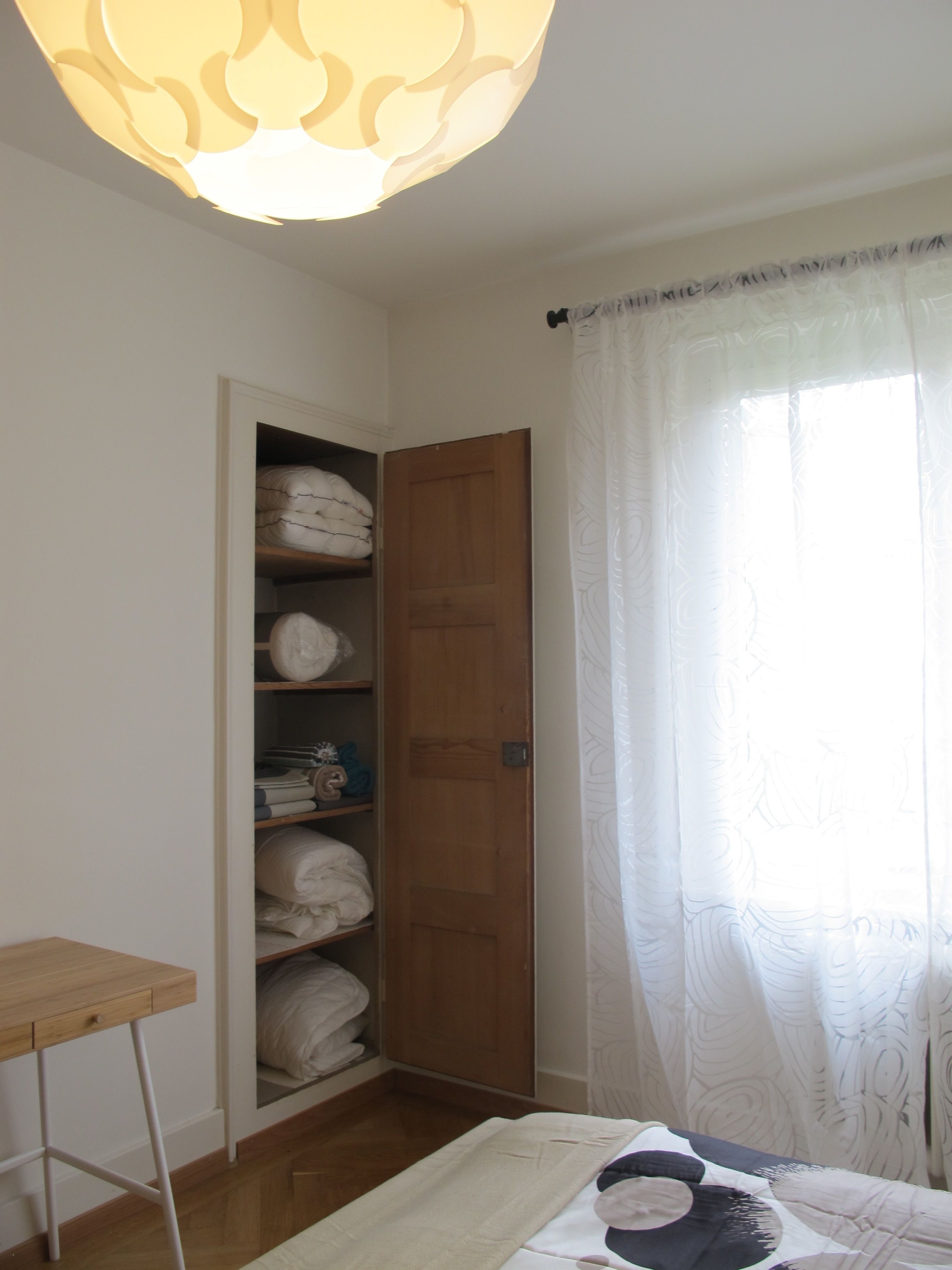 A bedroom with a wooden wardrobe, a bedside table, and a window with sheer curtains.