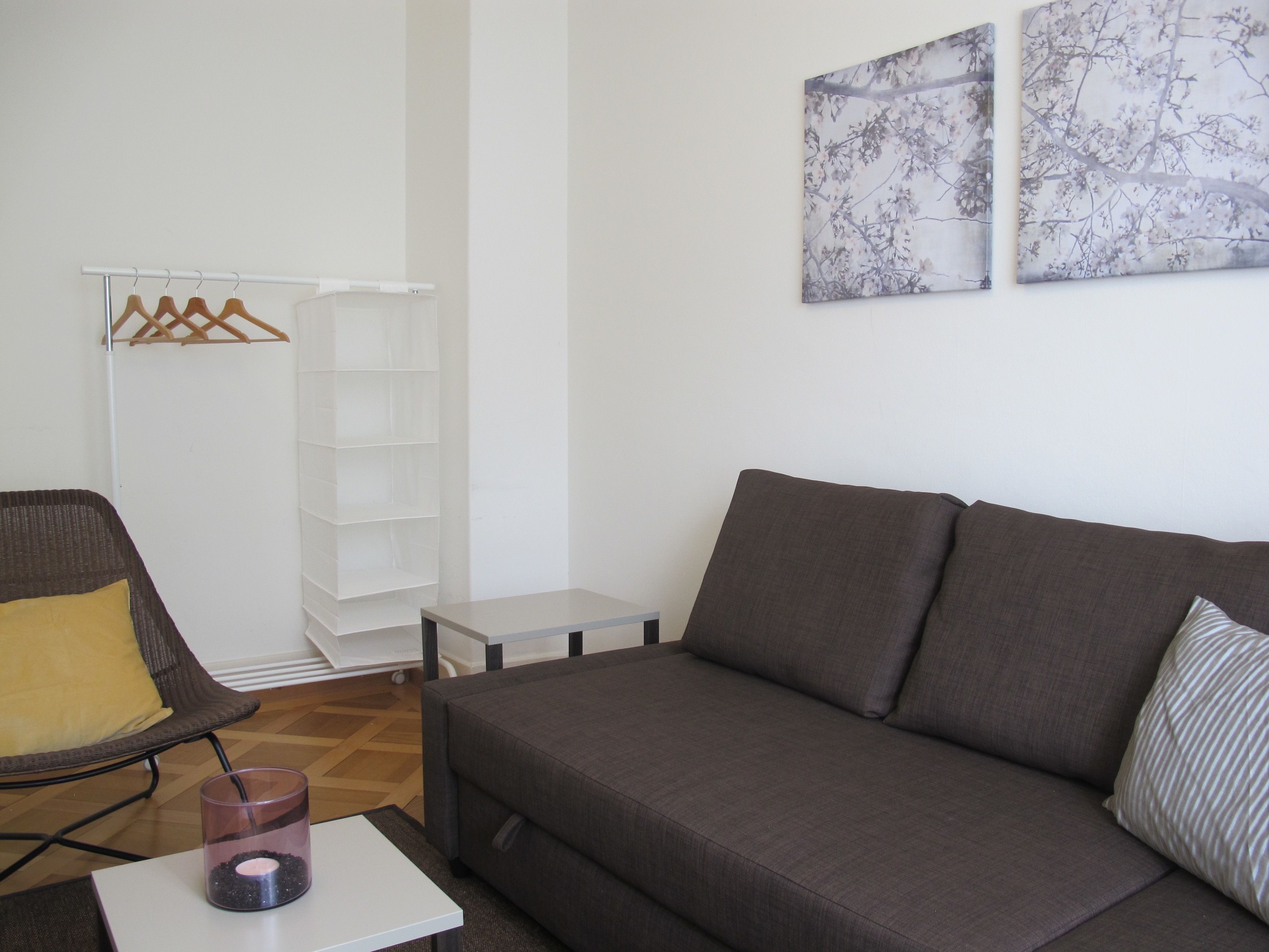 Uma sala de estar moderna com um sofá cinzento, uma cadeira de vime com uma almofada amarela, uma pequena mesa branca e um castiçal de vidro.