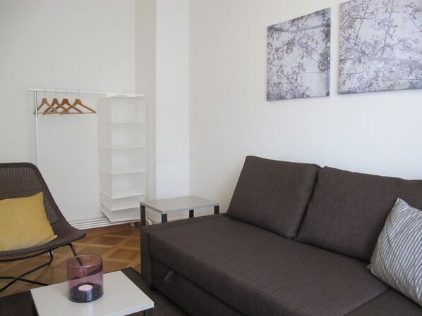 A modern living room with a grey sofa, a wicker chair with a yellow cushion, a small white table, and a glass candle holder. - Zurich Furnished Apartments (Zürich)