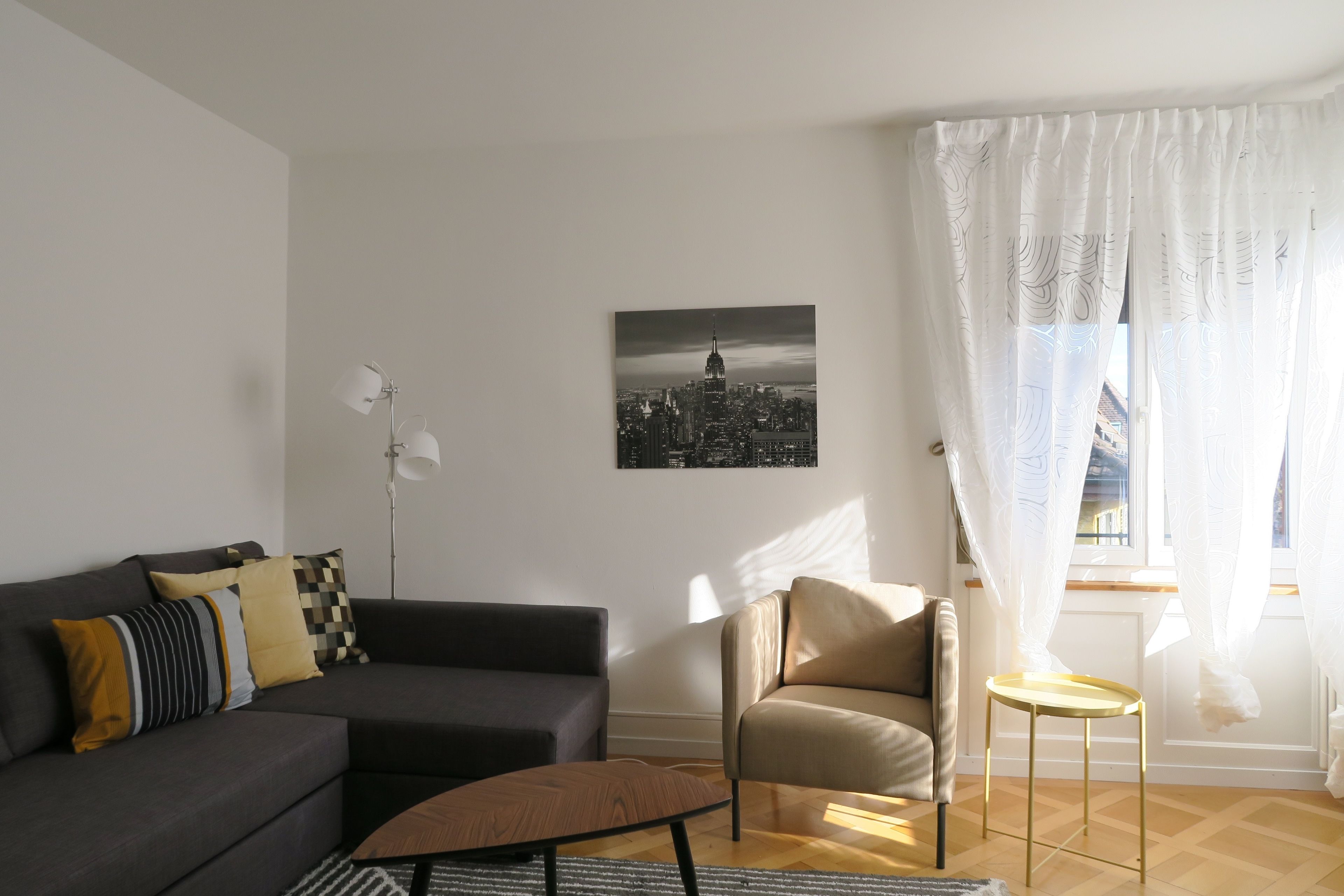 Ett modernt vardagsrum med en grå soffa, en brun pall, en beige fåtölj och ett litet runt bord. En svartvit målning som föreställer en stadsbild hänger på väggen.