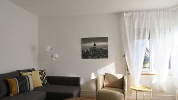 Ett modernt vardagsrum med en grå soffa, en brun pall, en beige fåtölj och ett litet runt bord. En svartvit målning som föreställer en stadsbild hänger på väggen.